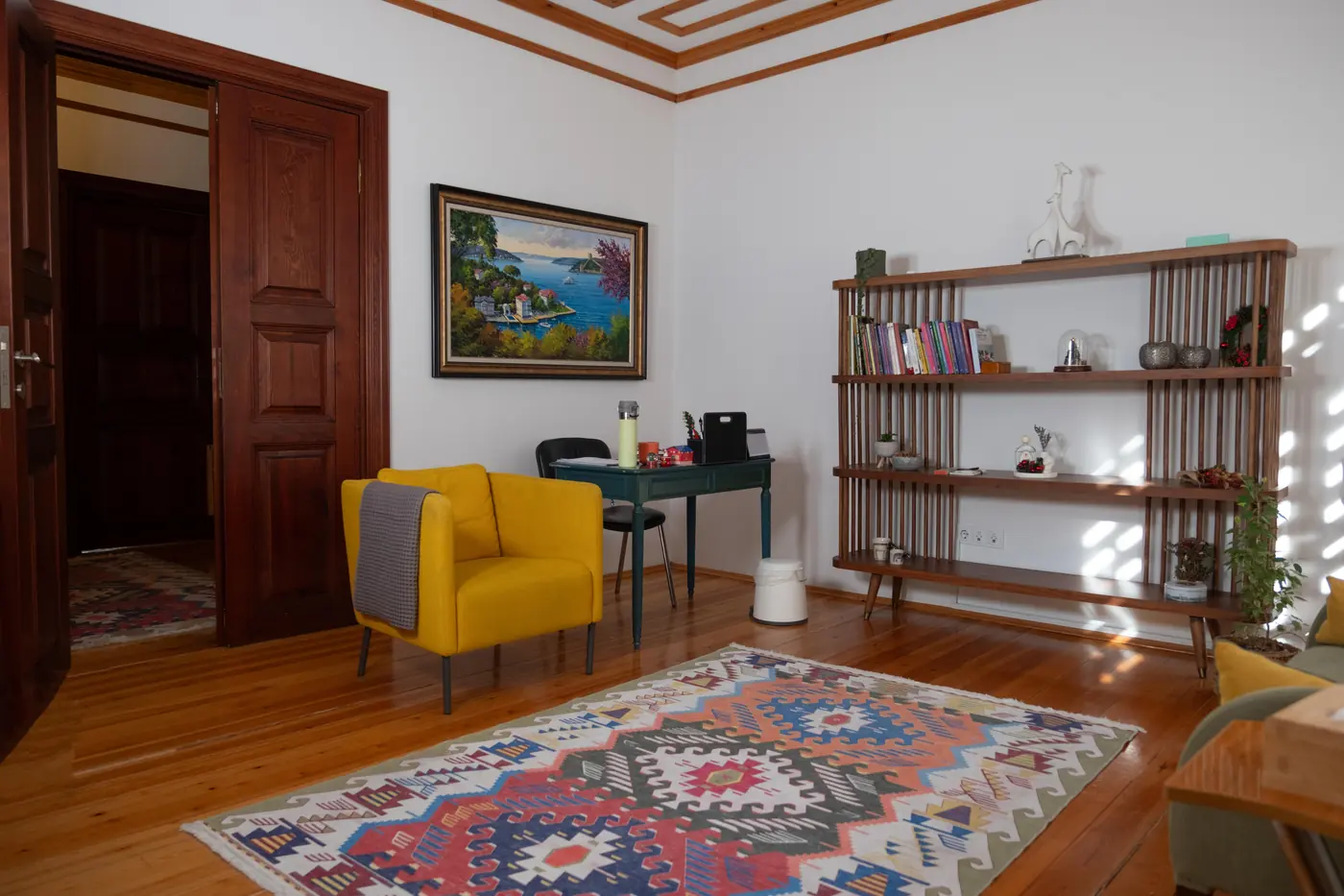 Kilim üzerinde sarı koltuk, ahşap raflar ve tablo bulunan terapi odası