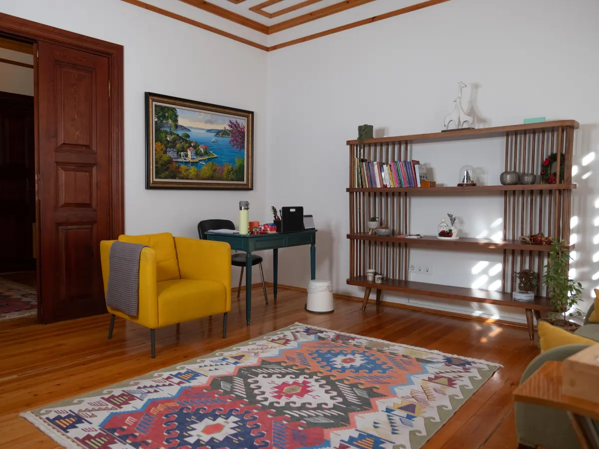 Kilim üzerinde sarı koltuk, ahşap raflar ve tablo bulunan terapi odası